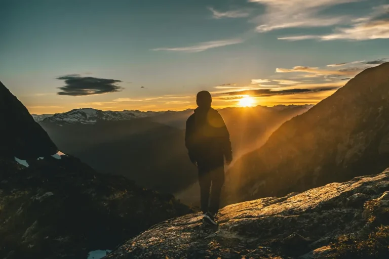 Silhouet van een wandelaar in jas die op een rotsige bergtop staat en richting een gouden zonsondergang loopt. Op de achtergrond een diep bergdal met besneeuwde toppen aan de horizon.