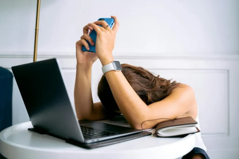 Een vrouw zit aan een bureau, hoofd voorover op een open laptop, haar beide handen houden een smartphone vlak boven haar hoofd. Beeld van informatie-overload en mentale uitputting door schermen.